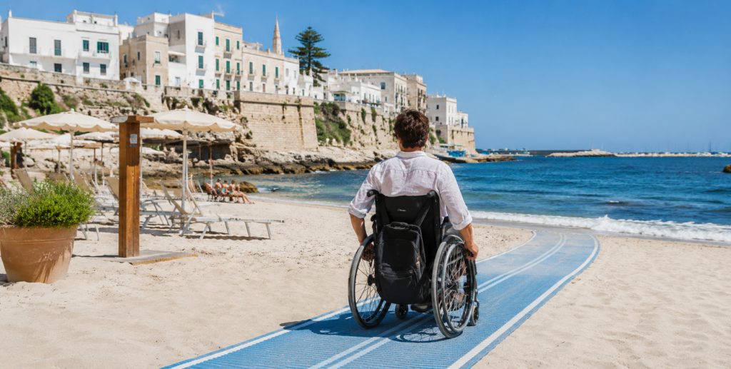 Turista in sedia a rotelle percorre un tappeto accessibile verso il mare su una spiaggia pugliese.